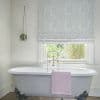 Patterned grey Roman blind, partially lowered with swirling texture, filtering daylight over a window above a freestanding claw-foot bathtub with chrome faucet, pink towel and hanging plant.