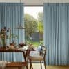 Floor-length pleated pale-blue curtains, drawn slightly apart to frame a central opening, revealing a sunlit dining table set for breakfast and a lush backyard garden beyond.