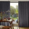Thick pleated dark-gray curtains, hung and drawn aside to reveal a glass door, framing and filtering daylight; a dining table set with pancakes and pendant lamp overlooks a backyard.