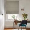 Beige-grey Roman blind, lowered with soft folds over a sash window; filtering daylight and obscuring the view; neutral study with wooden desk, teal chair, mirror and vase of flowers.