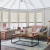 Beige pleated cellular blinds lowered mid-window, filtering daylight; across a bay of tall windows beneath a glazed roof, framing a bright conservatory living room with sofa, coffee table and blue rug.