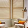 Horizontal beige venetian blinds covering bay windows, tilted closed to softly filter warm daylight; in a cozy living-room corner with textured boucle armchairs, orange cushions, a floor lamp, and neutral walls.