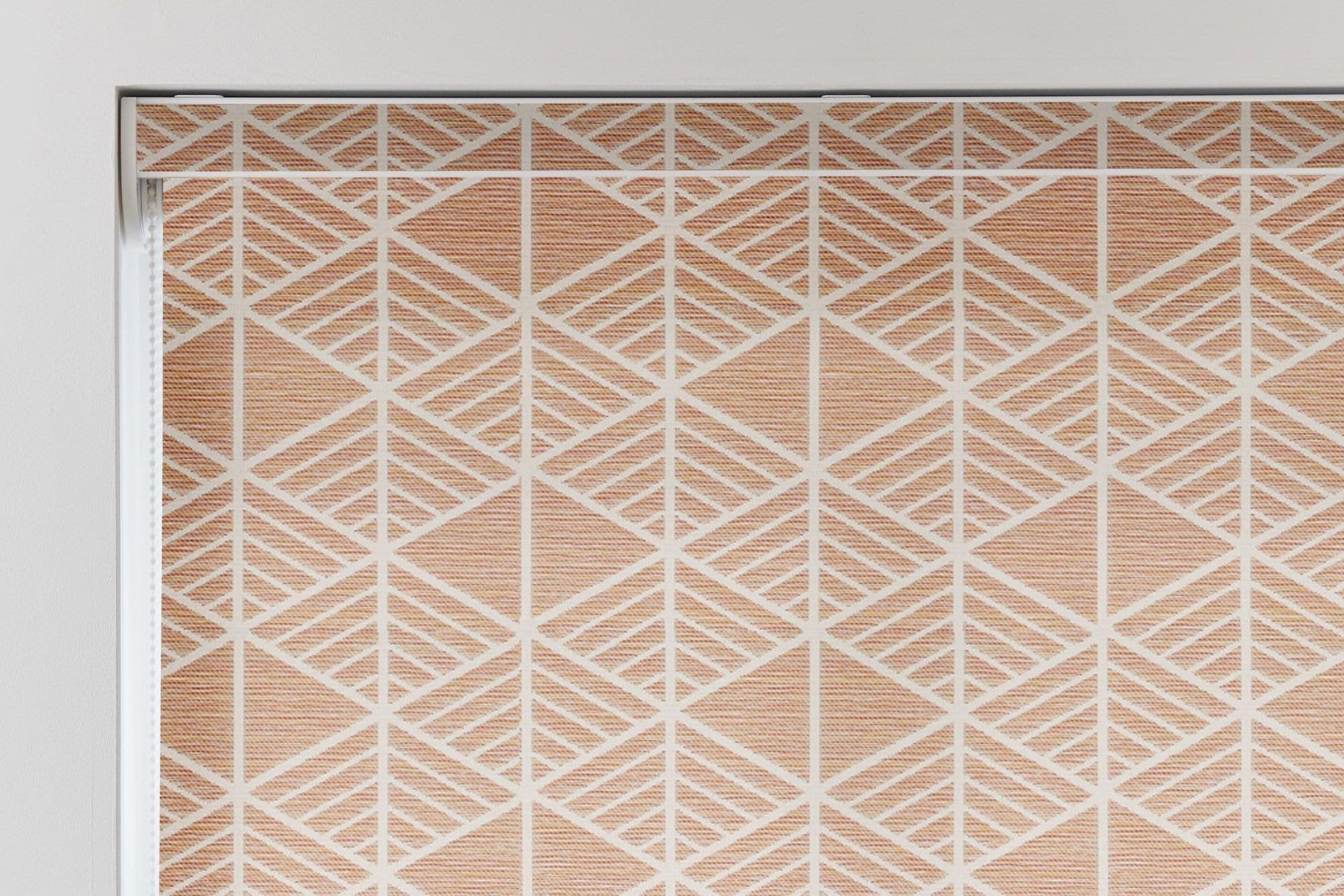 A closed roller blind with a peach-and-white geometric diamond pattern covers a window; it sits within a white frame, filtering soft indoor light in a neutral interior.