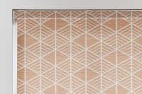 A closed roller blind with a peach-and-white geometric diamond pattern covers a window; it sits within a white frame, filtering soft indoor light in a neutral interior.