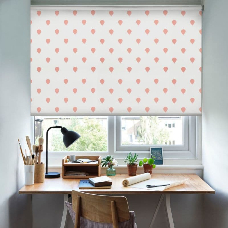 A white roller blind printed with small pink balloon motifs is lowered, filtering daylight over a home desk workspace with a lamp, art brushes, books and potted plants.