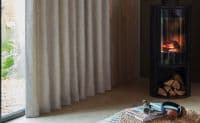 Creamy grey, floor-length pleated curtains closed over a sliding glass door, filtering soft daylight; living-room corner with a lit cylindrical wood-burning stove, stacked logs, woven pouf, magazines and headphones.
Contura