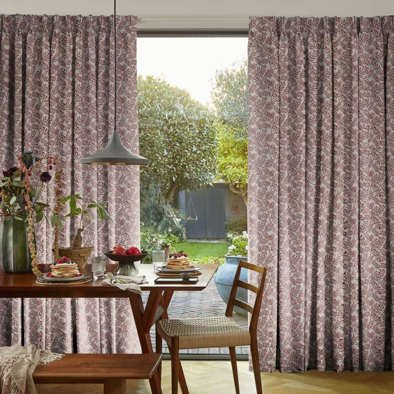 Pleated floor-length curtains with red leaf pattern, parted slightly, filtering daylight through a glass door; breakfast table, pendant lamp and wooden chairs in a dining room opening onto a garden.