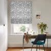 Roman blind with grey squiggle pattern, partially lowered and filtering daylight; over a window above a desk with vase of flowers, teal chair and framed print in a bright room.