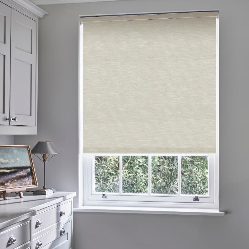 Beige textured roller blind lowered over a white sash window, covering upper panes and filtering daylight; lower panes reveal garden foliage; neutral gray room with white cabinetry, lamp, and artwork.