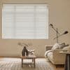 White horizontal Venetian blinds covering a large window, fully closed and filtering daylight; beige living room with sofa, wooden coffee table, patterned rug and slim black floor lamp.