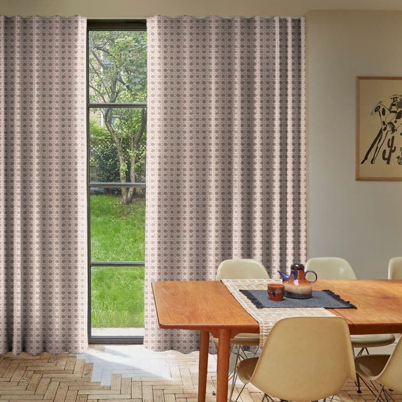 Floor-length pleated patterned curtains (pale pink geometric), drawn mostly closed with narrow central gap revealing a tall black-framed window; dining room setting with wooden table, beige chairs, green lawn outside.