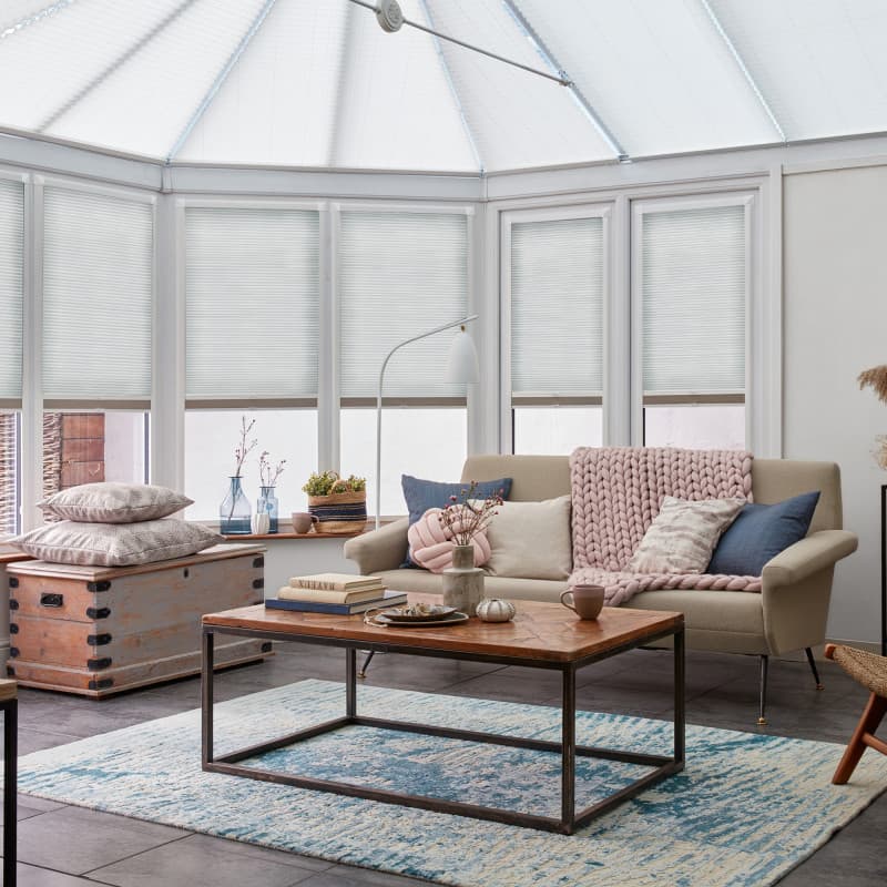 White honeycomb blinds covering tall conservatory windows and roof panels, lowered and closed, gently diffusing daylight; bright sitting area with beige sofa, knit throw, wooden coffee table, blue rug.