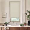 Beige roller blind lowered over a kitchen window above a stainless sink; countertop displays faucet, bowl of leafy greens, small potted plants and folded towel, warm wood cabinets below.