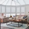 White cellular pleated blinds cover bay windows and roof panels, lowered to filter soft daylight; cozy living room with beige sofa, pink chunky throw, wooden coffee table and blue rug.