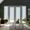 Patio glass doors framed by tall side windows; vertical blinds lowered halfway, filtering light. Context: modern living room with green armchairs, glass coffee table on chevron rug, plants and garden beyond.