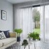 Sheer floor-to-ceiling grey curtains with soft pleats, drawn across a sliding glass door, gently filtering daylight; living room with sofa, patterned tile floor, potted plants, and a grassy backyard.