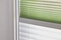 Pleated blinds—green upper and white lower—fitted inside a window frame; partially closed, filtering soft daylight; close-up view of a neutral interior wall and sill.