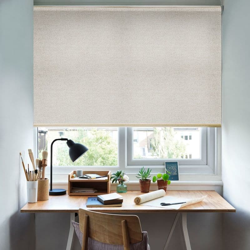 Beige textured roller blind lowered across the window, softly filtering daylight; small home desk beneath with a lamp, brushes, notebooks and potted succulents, bright residential view outside.