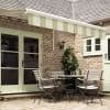 Striped green-and-white retractable awning extended from the roof, shading and softly filtering light over a brick-walled patio with mint-green French doors, a bistro table and chairs, and a potted climber.