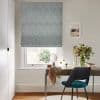 A textured grey Roman blind covers a window, lowered to filter daylight; a small desk with mirror, chair, vase of flowers and framed seaside print in a bright home office.