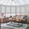 White pleated blinds fitted to the sloped conservatory roof and bay windows, lowered to filter soft daylight; cozy sunroom living area with beige sofa, wooden coffee table, and chunky pink throw.