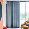 Floor-to-ceiling textured gray curtains with wave-top heading, mostly closed and filtering daylight from a large glass window; modern living room with mustard armchair and garden view.