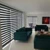 Horizontal zebra blinds on large corner windows, alternating opaque and sheer bands, filtering soft daylight into a modern living room with teal sofas, wooden coffee table, and a gray armchair.