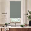 Gray roller blind covers a window above a sink and faucet, in a modern kitchen with wooden cabinets, potted plants, a bowl of vegetables, and a framed sketch on the wall.