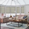 Beige pleated cellular blinds across six tall conservatory windows, lowered about three-quarters and filtering soft daylight; cozy sunroom with pitched translucent roof, neutral sofa, chunky pink throw, wooden coffee table.