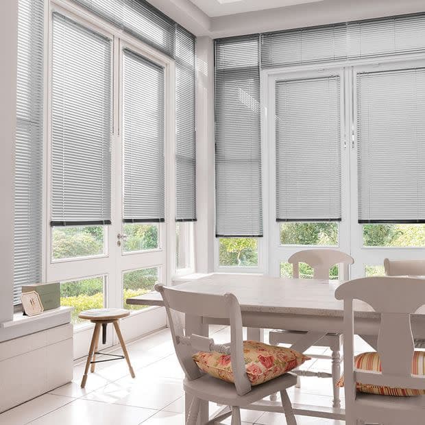 Grey horizontal Venetian blinds cover tall glass doors and windows, slats mostly closed and filtering daylight; bright white dining area with wooden table, chairs, floral cushions and garden view.
