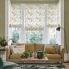 Three patterned roman shades (fern print) covering a three‑part bay window, partially lowered and filtering daylight; in a living room with beige sofa, cushions, coffee table, plants and floor lamp.