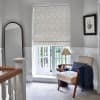 Patterned fabric Roman blind lowered over recessed window, subtle gray wave motif; filtering daylight into a bright upstairs landing with upholstered armchair, navy throw, floor lamp, and garden view.