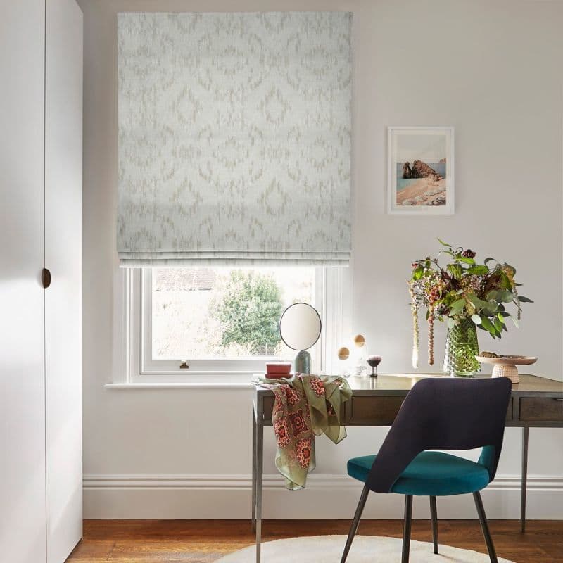 Pale patterned Roman blind lowered over a window, with subtle ikat-diamond fabric; filtering soft daylight onto a desk with mirror, vase of flowers and teal chair.
