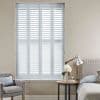 White full-height plantation shutters (four narrow panels) closed over the window, horizontal louvers slightly open and filtering bright daylight into a neutral-toned bedroom with armchair, bedside table, and lamp.
