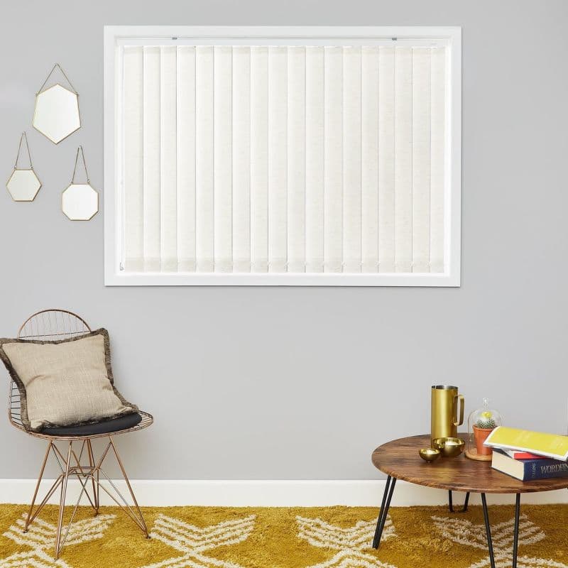 Cream vertical fabric blinds covering a centered window, closed and gently diffusing daylight; minimal living room with gray walls, wire chair with cushion, round wooden coffee table, mustard patterned rug.