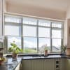 Kitchen sink and faucet sit beneath a large multi-pane window; sheer roller shades diffuse sunlight as potted plants and fruit bowls rest on granite countertops, framing a distant hillside view.