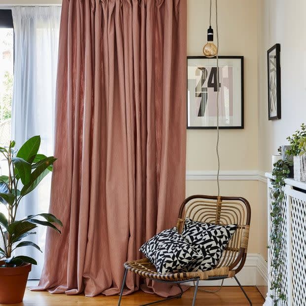 Dusty-rose floor-length curtains with sheer white voile behind, pleated and pooling; filtering daylight from a sliding door — corner living room with rattan chair, black-and-white cushions, plant, hanging bulb, framed 24 7