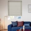 A beige cellular blind, lowered with horizontal ribs, filtering daylight over a sash window showing a roof and trees, in a living room with a blue sofa and lamp.