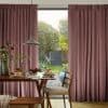 Pleated floor-to-ceiling dusty-rose curtains, hung with deep gathers and parted centrally, framing and filtering morning light onto a dining table set for breakfast, overlooking a small green garden with trees.