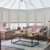 Pleated cellular blinds across tall conservatory windows and glazed roof, beige and fully lowered, diffusing soft daylight; a cozy sitting area with sofa, pink knitted throw, and wooden coffee table.