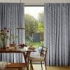 Floor-length grey curtains with dark leaf print, pleated and drawn aside; filtering daylight through sliding glass door. Dining table set for breakfast under pendant lamp, view of sunlit garden.