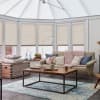 Beige pleated cellular blinds pulled down over tall conservatory windows, softly filtering daylight; cozy sunlit living room with sofa draped in a chunky pink knit, wooden coffee table, blue rug.