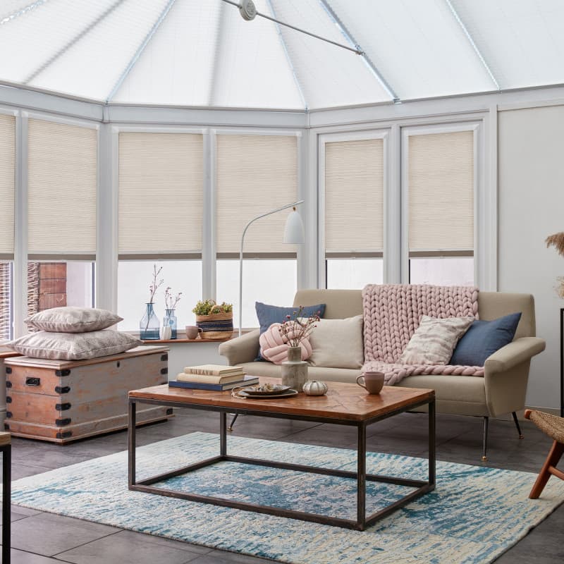 Beige pleated cellular blinds pulled down over tall conservatory windows, softly filtering daylight; cozy sunlit living room with sofa draped in a chunky pink knit, wooden coffee table, blue rug.