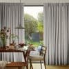 Full-length pleated gray curtains on a ceiling track, parted centrally—softly filtering daylight and framing a glass patio door—overlooking a dining table set for breakfast and a leafy backyard garden.
