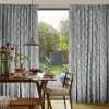 Gray textured floor-length curtains, hung to either side of a glass door, partially drawn and framing a sunlit garden; dining table set for breakfast, pendant lamp and wooden chairs inside.