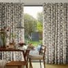 Floor-length pleated geometric-pattern curtains, parted centrally to reveal glass door, filter soft daylight; inside a wooden dining table set with pancakes and pendant lamp, outside a green garden view.