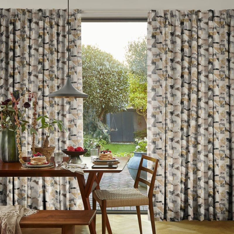 Floor-length pleated geometric-pattern curtains, parted centrally to reveal glass door, filter soft daylight; inside a wooden dining table set with pancakes and pendant lamp, outside a green garden view.
