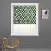 Roman blind with green geometric lattice, lowered to mid-window and filtering daylight; installed in a white frame on a gray wall, above a gold wire chair and a small abstract framed print.