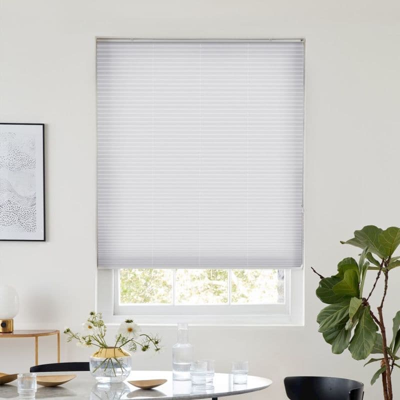White pleated cellular blind lowered, slightly raised at bottom, filtering daylight; minimal dining area with round table, vase of white flowers and glassware, framed art at left, fiddle‑leaf fig at right.