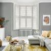 White plantation shutters covering a three-panel bay window, louvers tilted closed to diffuse soft daylight into a grey living room with radiator, sectional, yellow cushions, round coffee table and rug.
